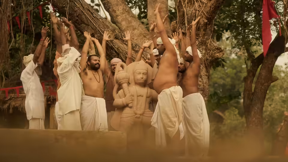 Men in white dhotis raise their arms around a large Hanuman statue beneath a tree