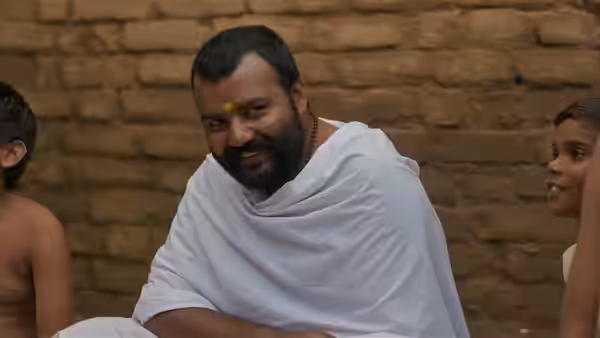 A smiling bearded man in a white wrap sits between two people against a brick wall