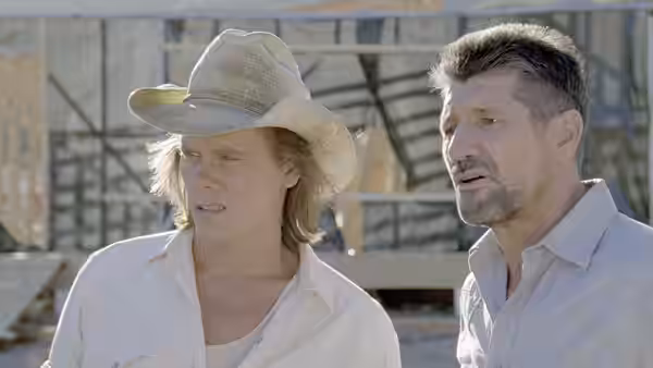 Rhonda LeBeck (Finn Carter) in a hat standing beside a man near buildings