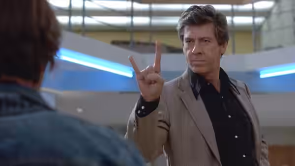 Richard Vernon holds up a peace sign while facing someone in the hallway