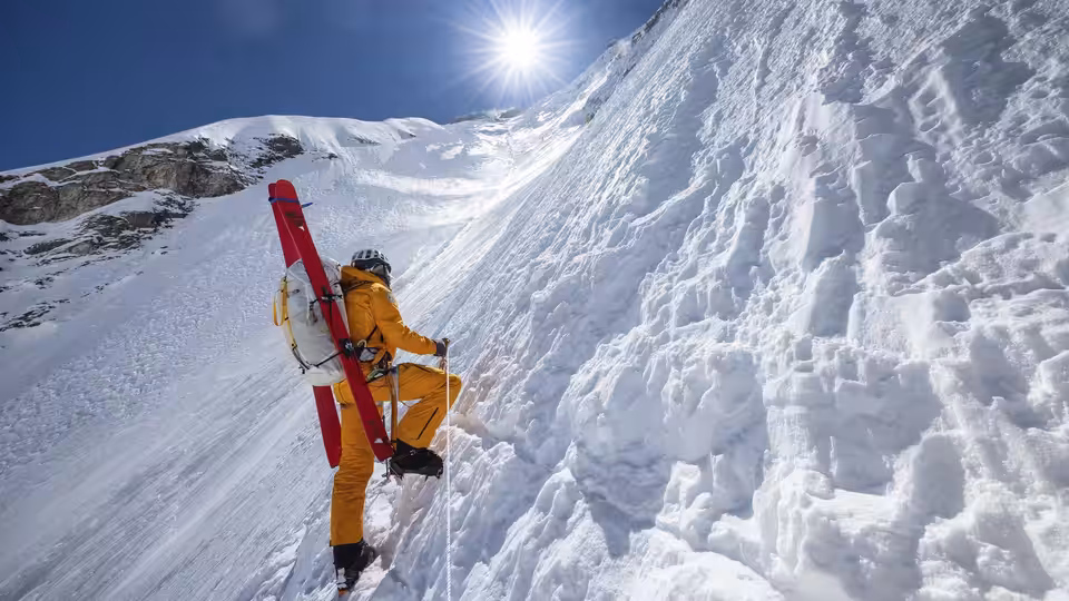A climber in an orange suit ascends a steep ice wall beneath the sun