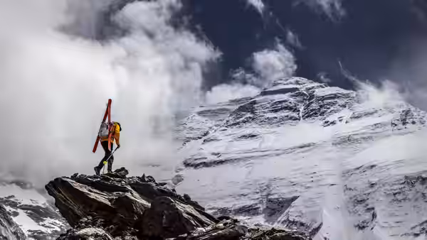 A climber stands on a rocky ledge above a snowy mountain slope