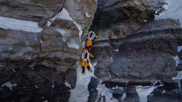 A climber in an orange suit scales a narrow crevice between dark rock walls