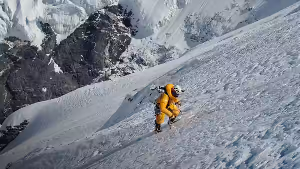 A climber in an orange suit traverses a steep snowfield below jagged cliffs