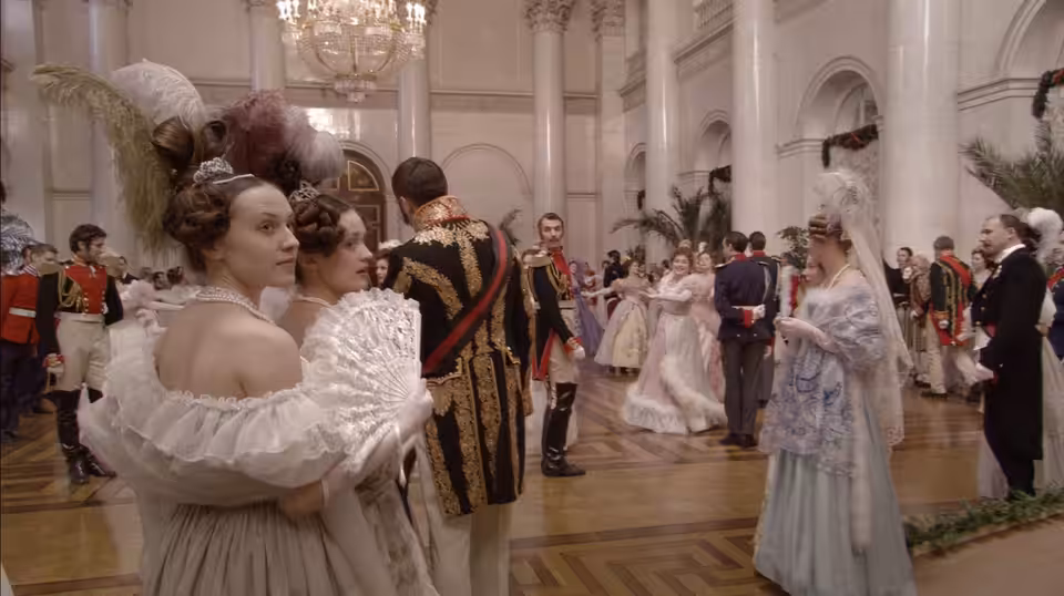 Guests in formal dress mingle and dance beneath chandeliers in a palace hall