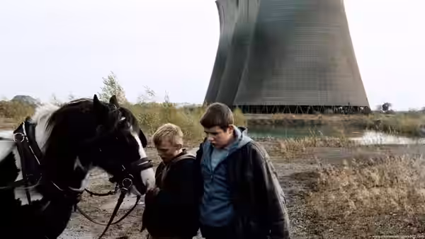 Two boys stand with a black and white horse before a cooling tower