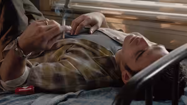 A young man lies on a bed as someone holds a cigarette above him