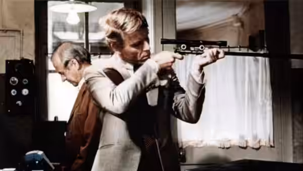 The Jackal (Edward Fox) aims a scoped rifle indoors while another man watches
