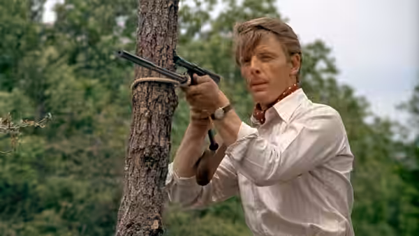 The Jackal (Edward Fox) holds a rifle beside a tree in the woods
