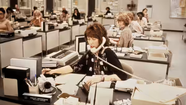 Violet Newstead (Lily Tomlin) answers a desk telephone amid rows of office workers and typewriters