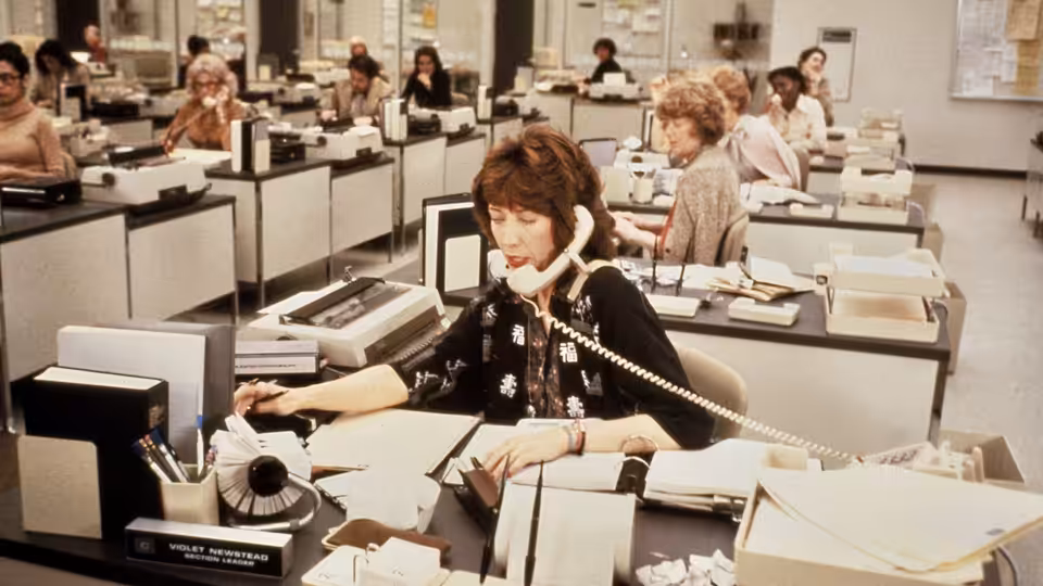 Violet Newstead (Lily Tomlin) answers a desk telephone amid rows of office workers and typewriters