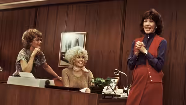 Judy Bernly (Jane Fonda) stands smiling beside Doralee Rhodes (Dolly Parton) and Violet Newstead (Lily Tomlin) at a desk