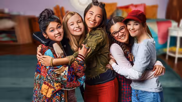 The girls hug together in a bedroom, smiling for the camera