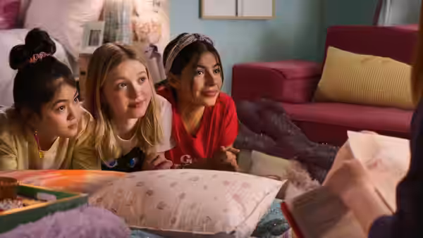 Three girls lean over bedroom pillows, watching something just beyond the frame