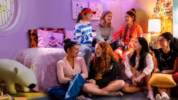 The girls gather around a bedroom bed, laughing and chatting together
