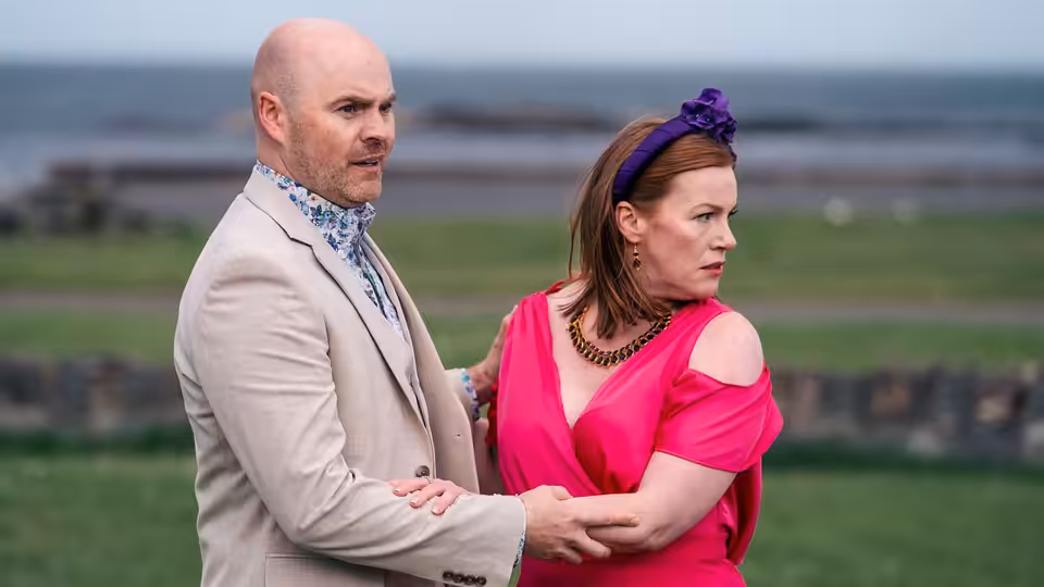 A man in a pale suit steadies a woman in pink as she looks away