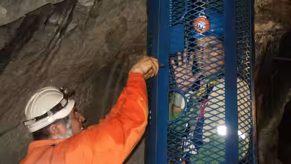 A helmeted miner in orange coveralls grips the mesh of a lift cage