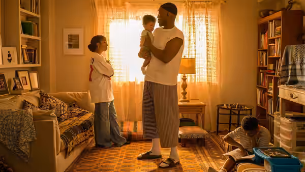 A man holds a toddler in a modest bedroom while a woman watches and a child sits nearby