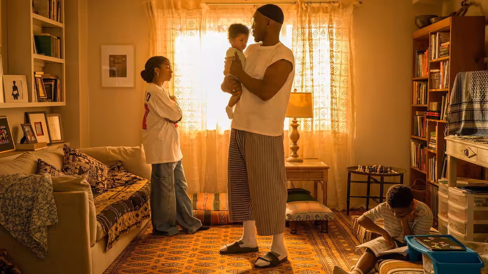A man holds a toddler in a modest bedroom while a woman watches and a child sits nearby