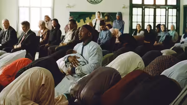 A man in a light robe sits among congregants seated in rows at a mosque