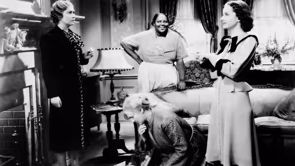 A seated older man bends forward as two women and a housekeeper stand in a living room
