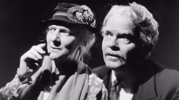 A woman in a decorated hat sits beside an older man in a suit