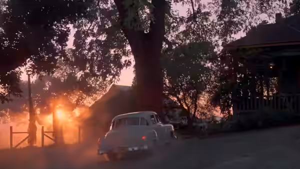 A vintage gray car driving away on a tree-lined road at dusk
