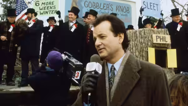 Phil Connors holding a microphone outdoors with crowd in top hats behind