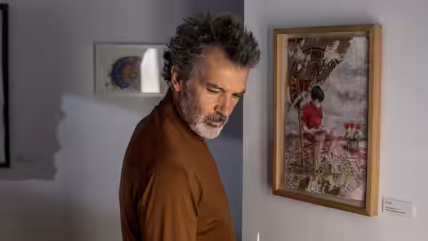 Man with curly graying hair and beard in brown turtleneck stands before a painting