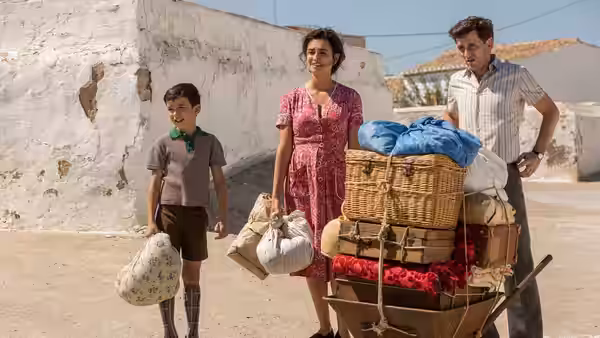 Woman in pink dress and young boy stand with a man beside a loaded luggage cart