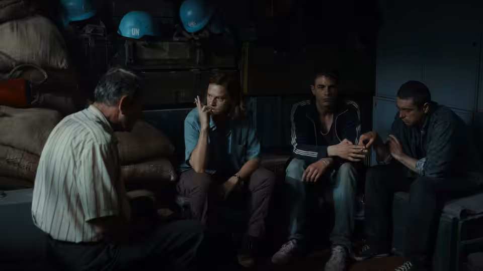 Aïda (Jasna Đuričić) talks with three men seated among sandbags and helmets