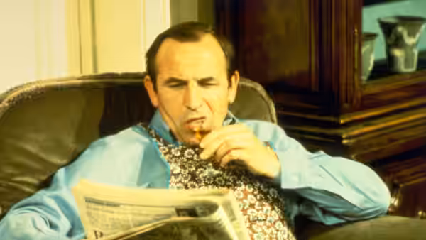 Reggie Perrin (Leonard Rossiter) reads a newspaper and eats while seated in an armchair