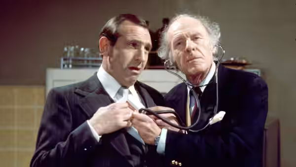 Reggie Perrin (Leonard Rossiter) stands as an older man listens to his chest with a stethoscope