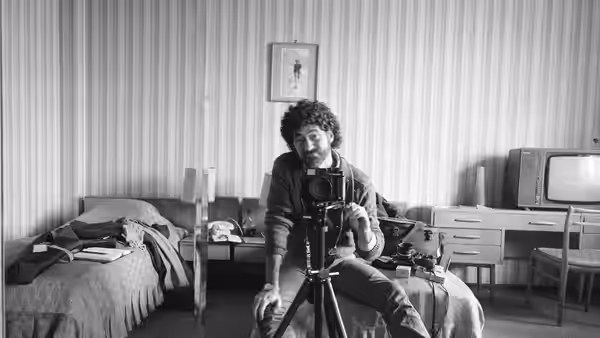 A man sits behind a camera on a tripod in a bedroom