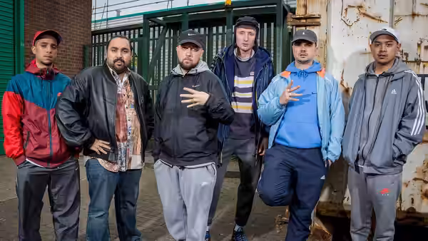 six men stand outside an industrial building near a green gate and fence