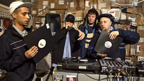 four men stand around a DJ setup holding vinyl records in a cluttered room
