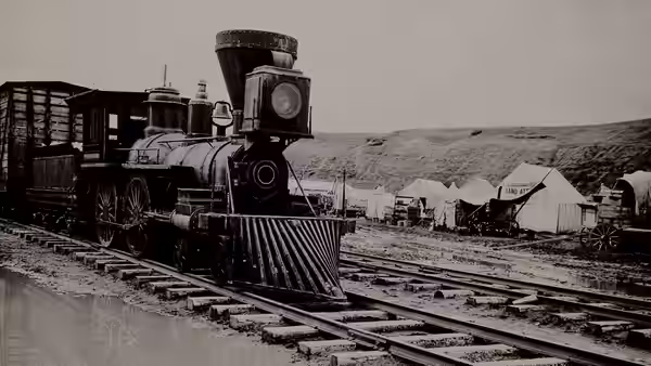 A steam locomotive travels along tracks past tents and railway buildings