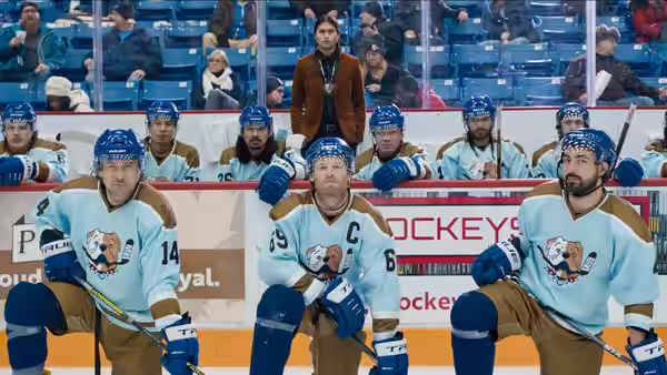 Hockey players sit on the bench as a man in a brown jacket stands behind them