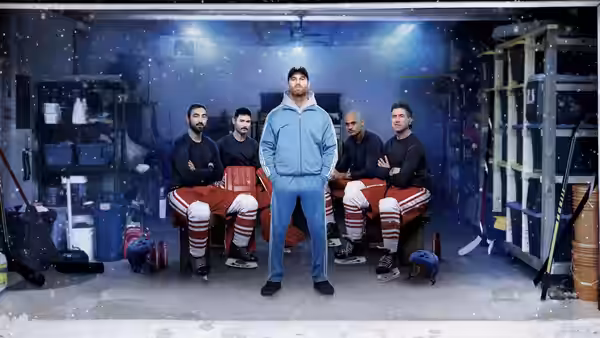 A man in a blue tracksuit stands before hockey players seated in a locker room