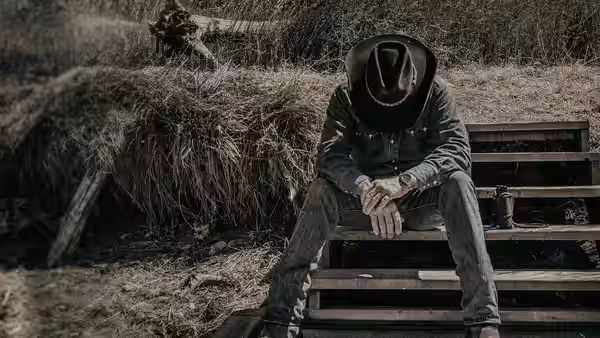 A man in a cowboy hat sits hunched on wooden bleachers beside a haystack