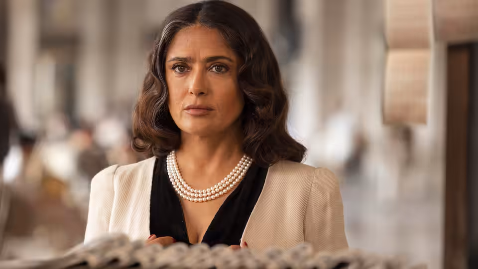 Nina (Salma Hayek) sits behind a desk in a beige jacket and pearl necklace