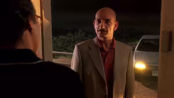 Dr. Roberto Miranda (Ben Kingsley) stands at a doorway beside a car with its headlights on