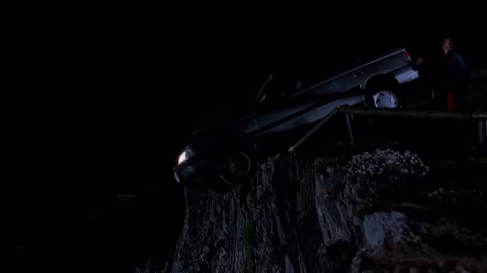 A car hangs nose-down over a rocky embankment as a figure stands nearby