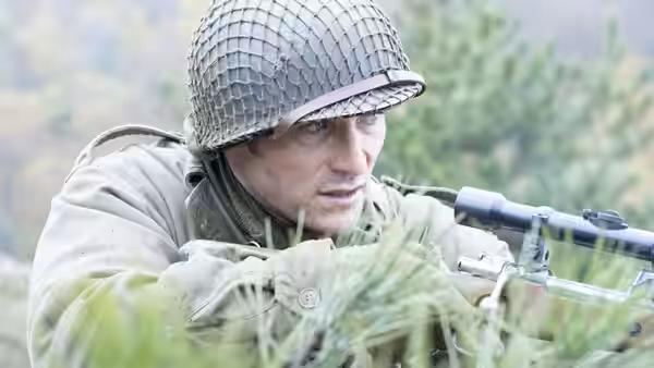 A helmeted soldier crouches in tall grass, aiming a rifle