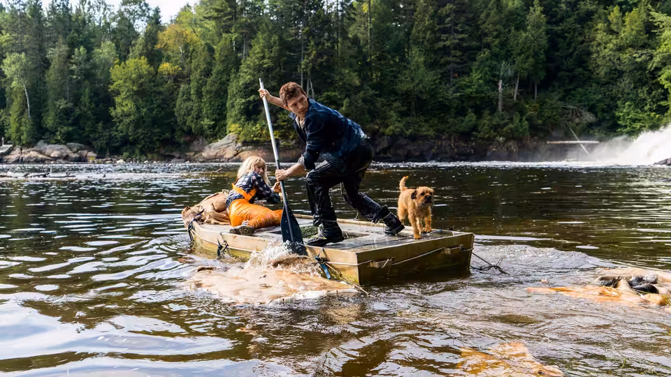 Todd Hewitt (Tom Holland) stands aboard a small boat beside a seated passenger and a dog