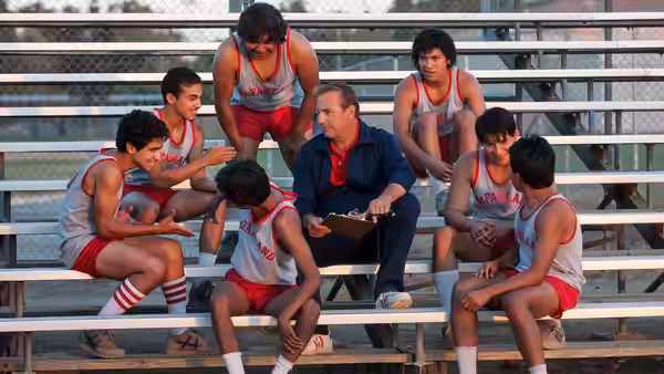 Jim White (Kevin Costner) reviews a clipboard with McFarland runners seated around him