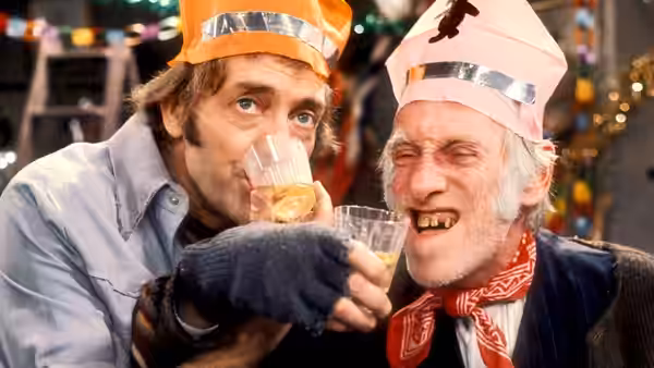 Harold (Harry H. Corbett) drinks from a glass next to Albert (Wilfrid Brambell) in party hats