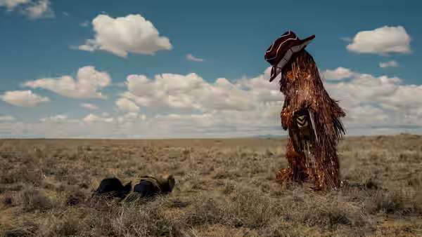 A bearded man in a tattered costume and hat stands in a field beside a prone figure