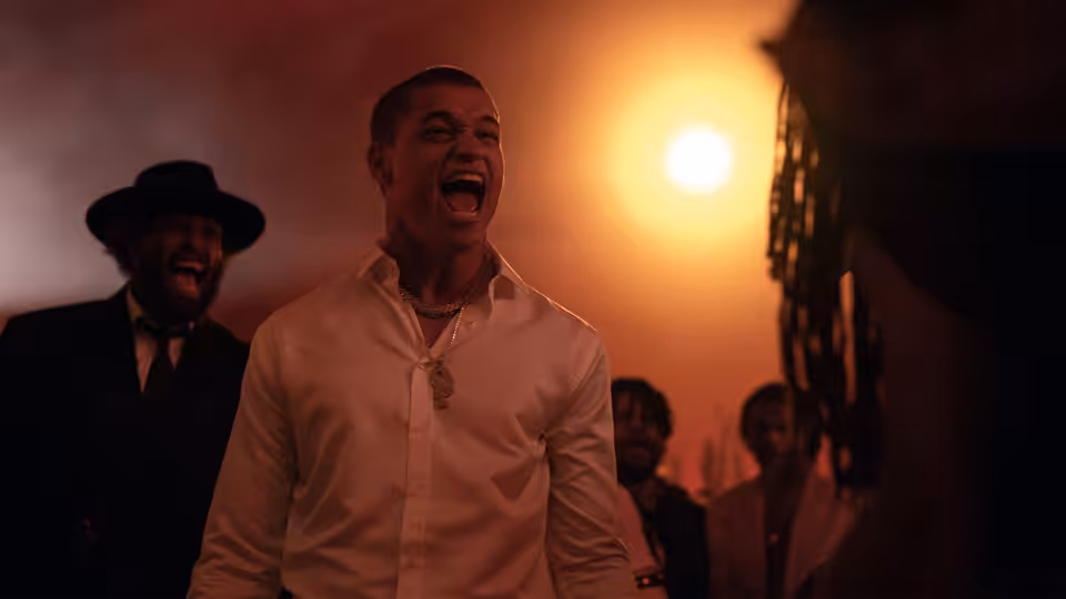 Cam (Tyriq Withers) laughs in a white shirt as men stand behind him at sunset