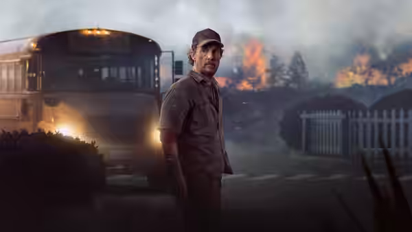 A uniformed man stands beside a school bus as wildfire burns behind him